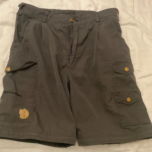 Grå shorts från Fjällräven - Snygga grå cargoshorts från Fjällräven med flera praktiska fickor och klassiska tryckknappar med logga. Shortsen är i slitstark bomull och har en loose passform, perfekta för sommardagar och äventyr utomhus. Storlek 46.