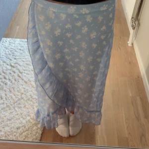 Blommig blå omlottkjol med volang - Supersöt ljusblå omlottkjol med vita blommor och volangkant. Kjolen är lång och har ett luftigt, romantiskt fall som ger en riktigt somrig vibe. Perfekt för dig som gillar mjuka färger och detaljer som sticker ut. Aldrig använt. Skriv för mer bilder!!