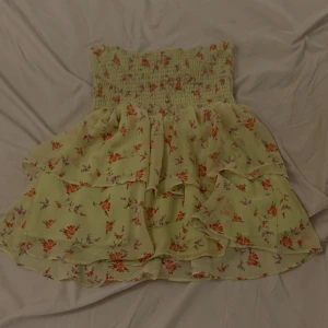 Blommig volangkjol från C i XS - Supersöt kort kjol från C i storlek XS. Kjolen är ljusgul med blommigt mönster i orange och lila, har smock upptill och flera lager volanger som ger en flowy känsla. Perfekt för dig som gillar romantisk och feminin stil.