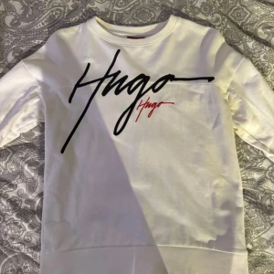 Vit sweatshirt från Hugo med print - Vit sweatshirt från Hugo med stor svart och röd logotyp på bröstet. Tröjan har rund hals, långa ärmar och är gjord av 100% ekologisk bomull. Snygg och clean design som passar perfekt till jeans eller joggers.