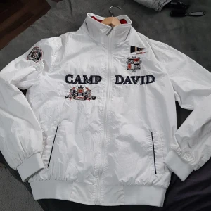 Vit vindjacka från Camp David L - Snygg vit vindjacka från Camp David med broderade detaljer och loggor på bröstet, ryggen och ärmen. Jackan har hög krage, dragkedja framtill och muddar vid ärmslut och midja. Perfekt för dig som gillar sportig och clean stil. På högerarmsmanget lite smutsig, sista bilden.