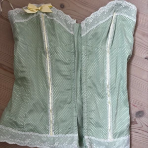 Mintgrön korsett från River Island - Superfin mintgrön korsett med vita prickar och spetsdetaljer från River Island. Toppen har en söt gul rosett upptill och är axelbandslös med omlott-design framtill. Materialet är mjukt och känns lyxigt, perfekt för att ge din outfit en extra touch. Passar i storlek small och xs
