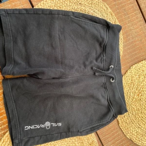 Svarta shorts från Sail Racing - Svarta shorts från Sail Racing med mjuk bomullskänsla och ribbad midja med snörning. De har fickor fram och en bakficka, samt Sail Racing-logga tryckt på ena benet.