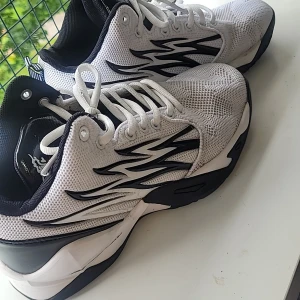 Vita och svarta sneakers med grafiskt mönster - Snygga vita sneakers med svarta detaljer och grafiskt mönster på sidorna. Ovandelen är i mesh för bra andningsförmåga och sulan är svart med coolt mönster undertill. Skorna har snörning och svart/vit plös med tryckt logga. Perfekta för dig som gillar sportig stil.