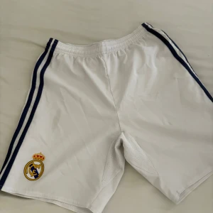 Vita Real Madrid shorts från Adidas - Snygga vita Real Madrid shorts från Adidas med blå ränder längs sidorna och klubbens emblem på ena benet. Tillverkade i lätt polyester med Climacool-teknologi för extra ventilation. Perfekta för fotbollsträning eller match. Elastisk midja för skön passform.