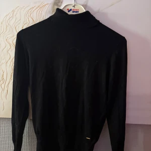 Svart polotröja Holly & Whyte M - Svart polotröja från Holly & Whyte i storlek M. Tröjan har lång ärm med snygga knappar vid ärmsluten och en klassisk polokrage. Perfekt att styla med jeans eller kjol för en clean look.