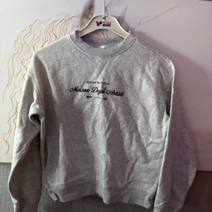 Grå sweatshirt Maison Degli Arrivi H&M XS - Grå sweatshirt från H&M i storlek XS med svart tryck 'Maison Degli Arrivi' framtill. Klassisk rund halsringning, ribbade muddar och mjukt material. Perfekt för en chill och stilren look.