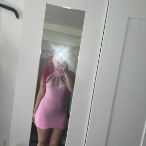 Rosa kortklänning med paljetter  - Säljer en rosa kortklänning från SHARE med tight passform. Klänningen är ärmlös och har en hög hals med silvriga paljettdetaljer framtill. Materialet känns mjukt och stretchigt, troligen bomull eller polyester. Perfekt för dig som gillar att sticka ut med färg och glitter. Lite lösa trådar under armen men inget som påverkar plagget 