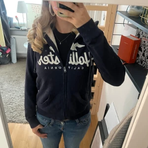 Mysig teddykofta från Hollister  - Säljer en mörkblå teddykofta från Hollister med vit fluffig insida och dragkedja. Kofta har broderad logga framtill, huva och två fickor. Står XS men skulle också kunna passa som S beroende på. Den har en liten fläck på insidan av koftan men som säkert går bort i tvätten!