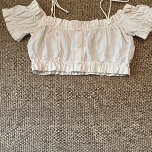 Vit croppad blus med volang - Supersöt vit croppad blus med offshoulder och korta puffärmar. Blusen har volangdetaljer, knappar framtill och knytband vid axlarna. Materialet är lätt och krispigt, perfekt för varma dagar. Passar dig som gillar romantisk och trendig stil.