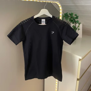 Svart Gymshark t-shirt XS - Svart t-shirt från Gymshark i storlek XS. Snygga meshdetaljer på axlarna och ventilerande partier. Tight passform och diskret logga framtill. Perfekt för dig som vill ha en sportig och stilren look på gymmet.