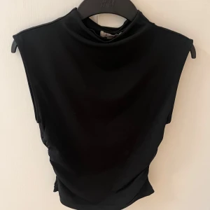 Svart polotopp från H&M - Svart topp från H&M med ärmlös design. Sitter tight och har snygga rynkade detaljer på sidorna som ger en cool siluett. Materialet är mjukt och stretchigt. Enkel och stilren look som funkar året runt och till fest