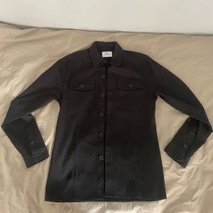 Svart overshirt från Ciszere - Stilren svart overshirt från Ciszere med klassisk krage och två bröstfickor med lock och knappar. Skjortan har långa ärmar med manschett och knäppning framtill. Perfekt att bära som lager över en t-shirt.