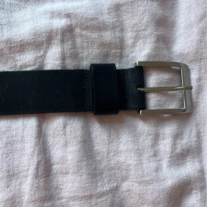 Svart klassiskt skärp i skinn - Snyggt svart bälte i äkta skinn med stilren silverfärgad metallspänne. Bältet är brett och har en enkel, tidlös design som passar till jeans eller kostymbyxor. Perfekt accessoar för att lyfta din outfit.