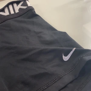 Svarta Nike Pro träningsshorts - Nike Pro shorts i svart med bred elastisk midja där loggan syns tydligt. Swoosh-detalj på benet. Sitter tight och är gjorda i ett stretchigt syntetmaterial som andas, perfekt för gym eller sport. Stilren design med platta sömmar för extra komfort. Defekt på bild 2 då sömnen gått upp lite, men inget större som märks.