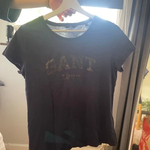 Mörkblå GANT t-shirt med tryck - Snygg mörkblå t-shirt från GANT med klassiskt tryck framtill. T-shirten har rund halsringning och korta ärmar, perfekt för en avslappnad och stilren look. Skön och mjuk känsla, passar till det mesta.