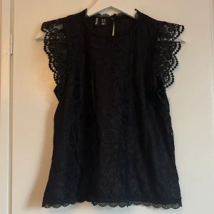 Svart spetsblus med volangärm  - Supersnygg svart blus från Pieces med spetsdetaljer och volangärmar. Blusen är ärmlös med vackert mönstrad spets längs axlar och nederkant. Perfekt för dig som vill ha en feminin och trendig look med extra detaljer.