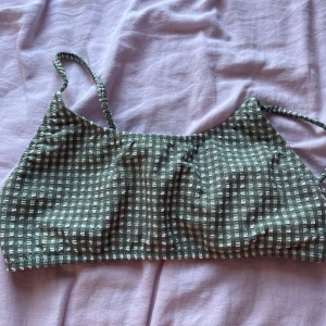 Grön rutig bikiniöverdel med spänne - Superfin bikiniöverdel i grönt och vitt med rutigt mönster och smala axelband. Toppen har ett snyggt metallspänne bak och vadderade kupor för extra komfort. Materialet är stretchigt och känns mjukt mot huden – perfekt för strandhäng eller pool.