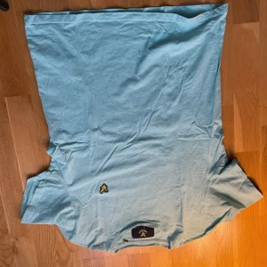 Ljusblå t-shirt från Lyle & Scott - Fräsch ljusblå t-shirt från Lyle & Scott med klassisk gul broderad fågel på bröstet. T-shirten har rund hals och korta ärmar, perfekt för chill dagar. Mjuk och skön bomull som känns nice mot huden. Snygg och enkel stil som funkar till jeans eller shorts.
