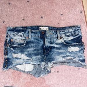 Blå jeansshorts med slitningar - Snygga blå jeansshorts från Perfect Jeans med råa, fransiga kanter och slitningar framtill och baktill. Shortsen har nitar längs sidorna, klassiska fickor och knappstängning. Perfekt för dig som gillar en edgy och avslappnad stil.