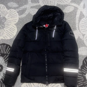 Svart pufferjacka Zavetti Canada M - Svart pufferjacka från Zavetti Canada med huva och reflekterande detaljer på ärmarna. Jackan har dragkedja framtill, bröstficka med dragkedja och broderad logga på ena ärmen. Perfekt för kalla dagar och riktigt snygg streetstil. Dene helt ny haft den bara 1 månad, priset kan minska max 100