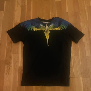 Marcelo Burlon svart t-shirt med vingar - Svart t-shirt från Marcelo Burlon County of Milan med färggrafiskt tryck av vingar i blått, grönt och gult över axlar och hals. Klassisk rund hals och korta ärmar. Materialet är mjuk bomull och passformen är normal. Perfekt statement-plagg för dig som gillar streetwear.