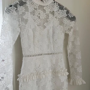 Vit spetsklänning med volanger Boohoo - Supersöt vit spetsklänning från Boohoo med blommigt mönster, långärmad och högt halsad. Volangdetaljer vid midjan och ärmslut ger en extra girly vibe. Perfekt för student eller sommarens alla tillfällen. Figurnära passform och transparenta partier. En jätte snygg studentklänning. Använt endast 2 gånger.