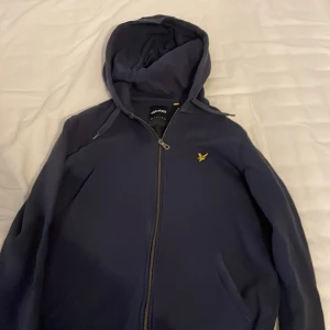 Marinblå hoodie från Lyle & Scott - Säljer en marinblå hoodie från Lyle & Scott med gul logga på bröstet. Tröjan har dragkedja framtill, huva med snörning och två fickor. Perfekt för chill dagar och enkel att matcha med jeans eller joggers.