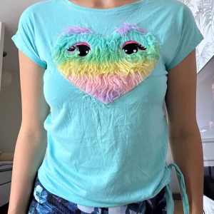 Turkos t-shirt med fluffigt hjärta - Turkos t-shirt med korta ärmar och knyt i sidan. På bröstet sitter ett stort fluffigt hjärta i regnbågsfärger med broderade ögon och rosa detaljer. Materialet är mjuk bomull och hjärtat har en pälsliknande yta som sticker ut. Perfekt för dig som gillar färg och roliga detaljer. Jag har VUXEN STORLEK S o den passar