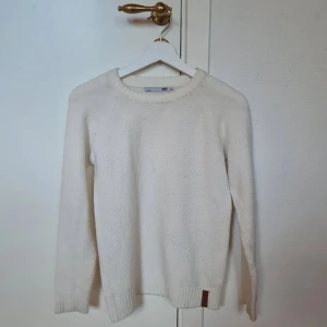 Vit stickad tröja från 157 - Mysig vit stickad tröja från 157 med rund hals och ribbade muddar vid ärmslut och nederkant. Tröjan har en enkel och clean design, perfekt för dig som gillar basic men ändå vill ha lite stil. Liten brun logodetalj vid nederkanten för extra touch.