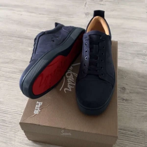 Mörkblå sneakers från Louboutin - Snygga mörkblå sneakers från Christian Louboutin i mocka med klassisk röd sula och broderad logga på hälen. Skorna har rund tå, snörning och en stilren design som sticker ut med den ikoniska röda undersidan.