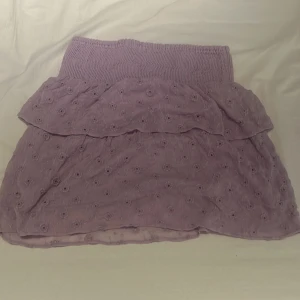 Ljuslila volangkjol med broderi - Supersöt ljuslila kortkjol med volanger och broderade blommor. Kjolen har en bred, smockad midja som sitter bekvämt och ger en snygg siluett. 