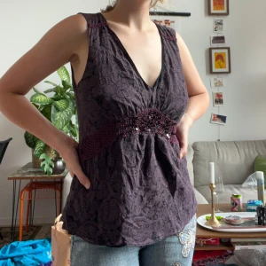 Lila blus med paljettdetalj från Stockh LM - Supersnygg lila blus med svart blommigt mönster och djup v-ringning. Midjan markeras av ett virkat band med paljetter som ger extra bling. Storleken är markerad som L men passar ändå ganska bra på mig som är S 