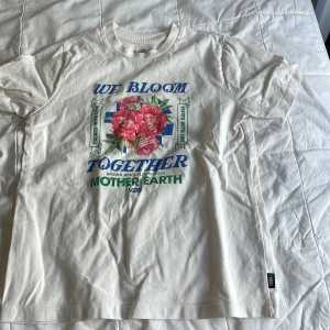 Vit blommig t-shirt från Vans - Vit t-shirt från Vans med tryck i blått och grönt samt stora rosa blommor på bröstet. Modellen är klassisk med rund hals och korta ärmar. Materialet är mjuk bomull och trycket har texten 'We Bloom Together' och 'Mother Earth'. Perfekt för dig som gillar coola prints.