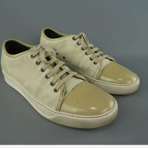 Beige lanvin sneakers med lackad tå - Snygga beige sneakers med snörning och rund tå. Skorna har en unik design med lackad tådel som ger extra glans och kontrast mot den matta textilen. Perfekta för dig som gillar neutrala färger och stilren look.