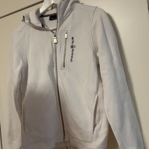 Vit hoodie från Sail Racing med dragkedja - Vit hoodie från Sail Racing med huva och hel dragkedja framtill. Tröjan har ett broderat Sail Racing-tryck på bröstet och praktiska fickor på sidorna. 