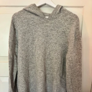Grå stickad hoodie från Ahlquist - Mjuk och chill grå stickad hoodie från Ahlquist Clothing i storlek L. Tröjan har huva, långa ärmar och ribbade muddar vid hals och ärmslut. Perfekt för kalla dagar när du vill vara både bekväm och snygg. Enkel design utan mönster eller detaljer.