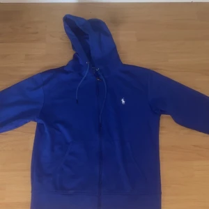 Blå hoodie från Polo Ralph Lauren - Snygg blå hoodie från Polo Ralph Lauren med klassisk vit broderad logga på bröstet. Tröjan har huva med snörning, dragkedja framtill och två fickor. Perfekt för chill dagar och streetstyle. Materialet är mjuk bomull som känns skönt mot huden.