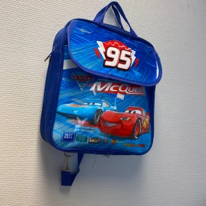 Blå Cars ryggsäck med McQueen-motiv - Säljer en blå ryggsäck med motiv från Disney Cars och Lightning McQueen. Väskan har justerbara axelband, bärhandtag och coolt tryck med nummer 95 och karaktärerna på framsidan. Perfekt för dig som älskar bilar och vill sticka ut i skolan. Helt ny 