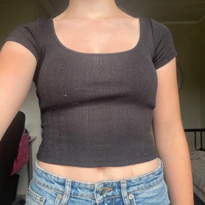 Svart croppad topp från H&M Divided - Svart croppad topp från H&M Divided med ribbad struktur och rundad, djup urringning. Kortärmad och figurnära passform som sitter snyggt till höga jeans eller kjol. Materialet är mjukt och stretchigt, perfekt för en chill och trendig look.