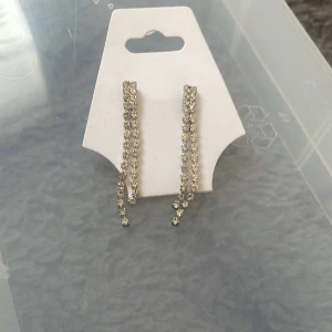 Glittriga silverfärgade örhängen - Snygga örhängen med två rader glittrande stenar i silverfärg. De är långa och hänger ner för en extra glamorös look. Perfekta för att ge din outfit lite extra bling. Stift med plastbaksida för bekväm användning.