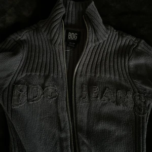 Mörkblå/grå ribbad kofta från BDG Jeans - Supersnygg kofta i en mörkgrå nyans med blå underton. Broderad BDG Jeans-logga framtill och dubbla dragkedjor. Hög krage och långa ärmar med fina detaljer vid muddarna.