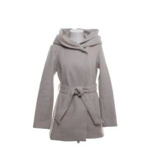 Ljusgrå kappa med huva från Amisu - Snygg ljusgrå kappa från Amisu med stor huva och knytskärp i midjan. Jackan har en stilren look med långa ärmar och markerad midja. Perfekt för dig som vill ha en clean och modern vibe under höst och vinter.