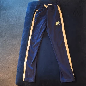 Nike Air blå mjukisbyxor med vita och svarta  ränder - Blå mjukisbyxor från Nike Air med vita och svarta sidoränder och logga på benet. Byxorna har resår i midjan och snörning för extra passform. Perfekta för chill eller träning, med en slim och sportig look. Finns jacka till, kika på mina annonser! 