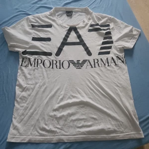 Vit EA7 Emporio Armani t-shirt - Snygg vit t-shirt från EA7 Emporio Armani med stor svart logga och tryck framtill. Klassisk rund hals och korta ärmar. Tillverkad i mjuk bomull som känns skön mot huden. Perfekt för dig som gillar stilrena och trendiga plagg med tydlig branding.