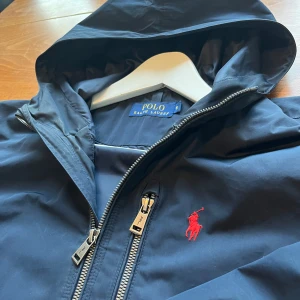  Ralph Lauren vindjacka - Snygg mörkblå vindjacka från Polo Ralph Lauren med huva och dragkedja fram. Klassisk röd Polo-logo på bröstet, lätt och skön. Bra passform, superfräsch och perfekt för regniga eller blåsiga dagar. Okej skick! 🟦🌬️💧