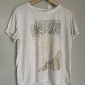 Vit t-shirt med paljettdetaljer Esprit - Vit t-shirt från Esprit med korta ärmar och ett unikt grafiskt tryck framtill i beige och grå toner. Paljettdetaljer ger extra glans och cool känsla. Tillverkad i mjuk bomull, noppror finns vid armhålor.