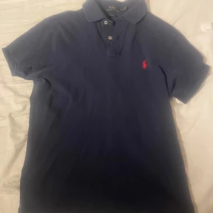 Mörkblå pikétröja från Ralph Lauren - Klassisk mörkblå pikétröja från Polo Ralph Lauren i bomull. Tröjan har korta ärmar, krage med knappar och den ikoniska röda broderade loggan på bröstet. Custom fit-modell som sitter snyggt och passar perfekt till jeans eller shorts.