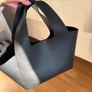 Svart shoppingväska i skinn Carin Wester  - Stilren svart shoppingväska med minimalistisk design och breda handtag. Väskan är rymlig och har en modern, kantig form som passar perfekt för att bära allt du behöver. Tillverkad i slätt skinn för en clean och tidlös look.