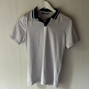 Vit piké från J.Lindeberg, storlek S - Snygg vit golf pikétröja från J.Lindeberg med klassisk krage och svarta/vita ränder. T-shirten har korta ärmar och en stilren design med diskret logga i nacken. Perfekt för en sportig och clean look.
